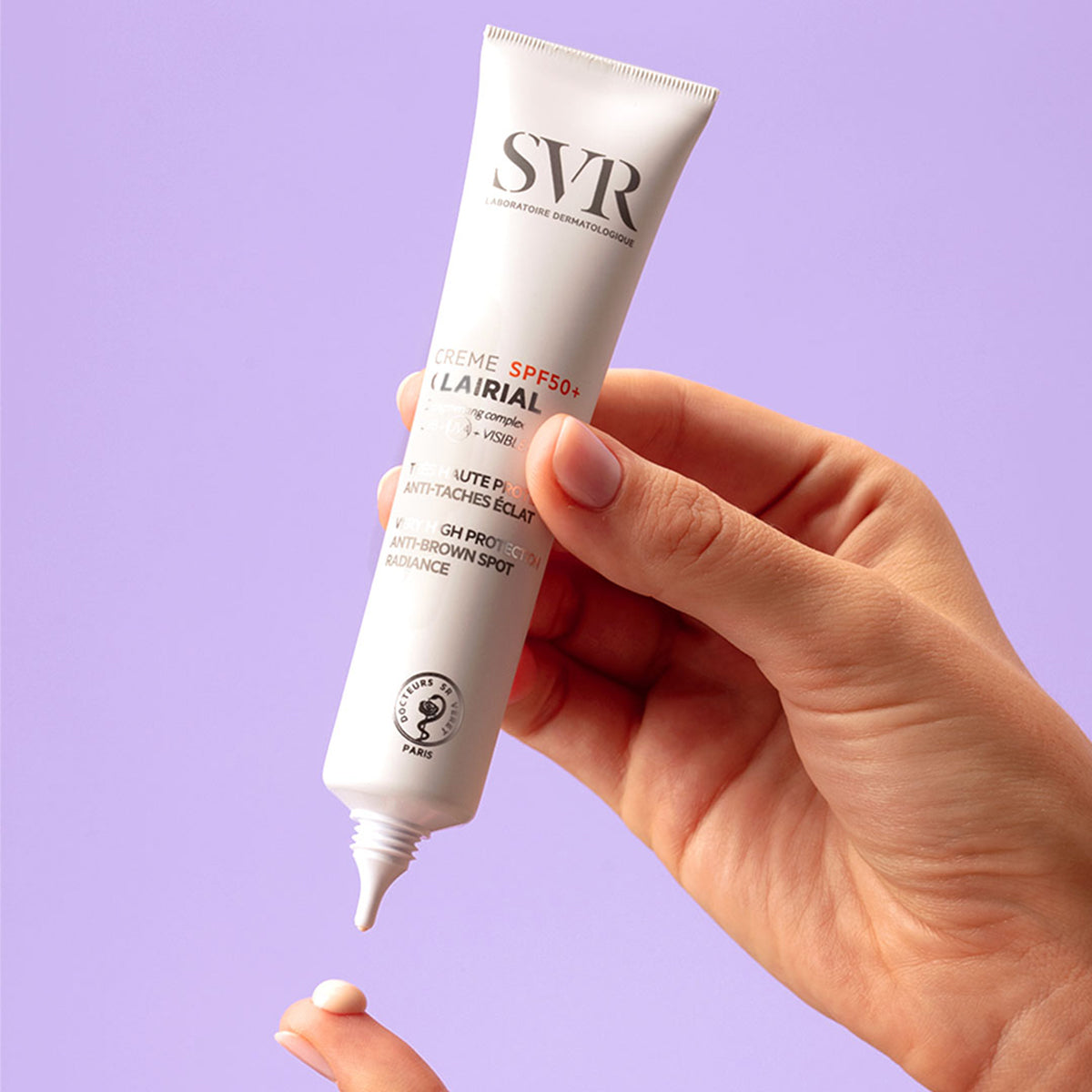Hand holding a white SVR Clairial SPF50+ cream tube dispensing a drop of product onto a finger against a purple background.