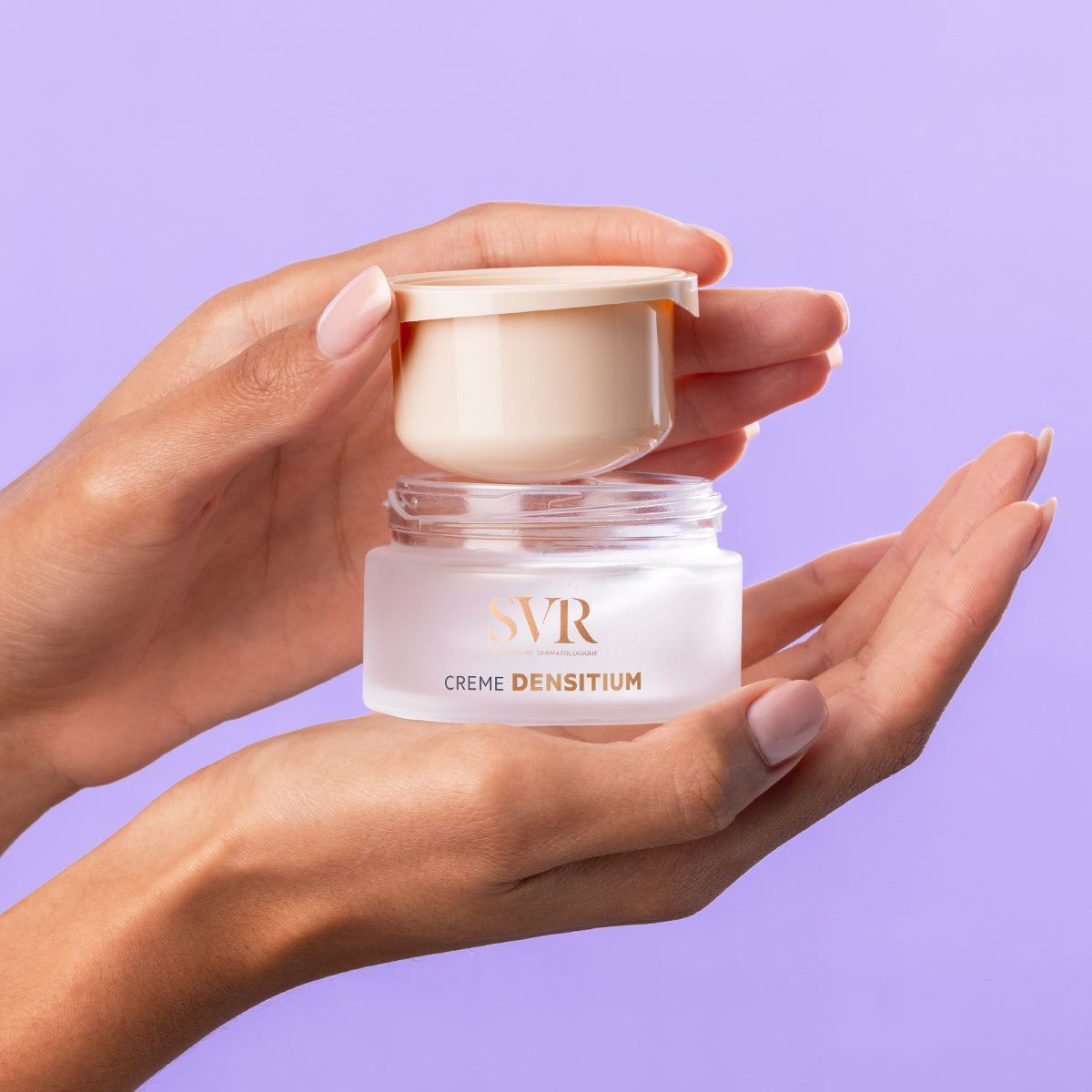 Hands holding an open SVR Densitium Creme jar with a frosted glass base and beige lid against a purple background.