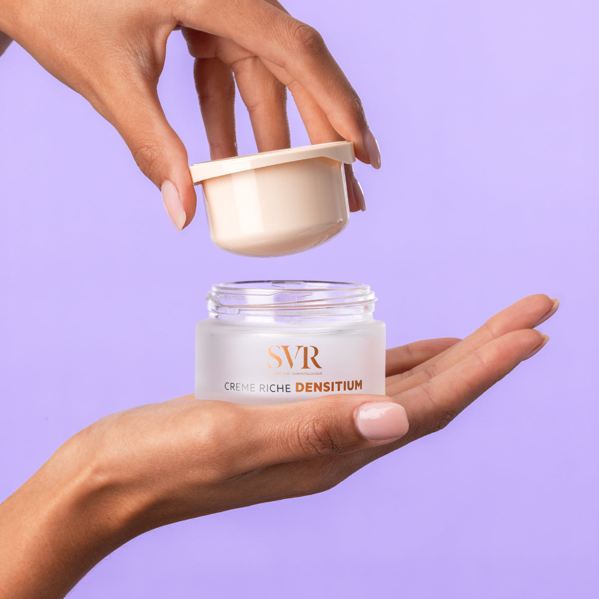 Hands holding an open jar of SVR Densitium Creme Riche with a frosted glass base and a pale pink lid against a purple background.