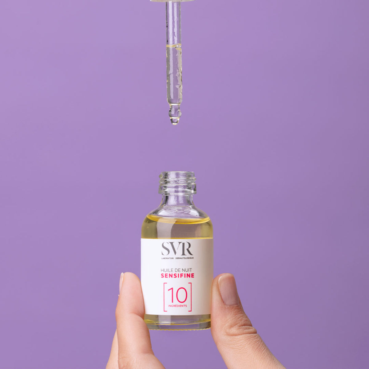 Frasco de vidrio de SVR Sensifine Huile de Nuit con gotero sobre fondo morado, mostrando el aceite facial reparador.