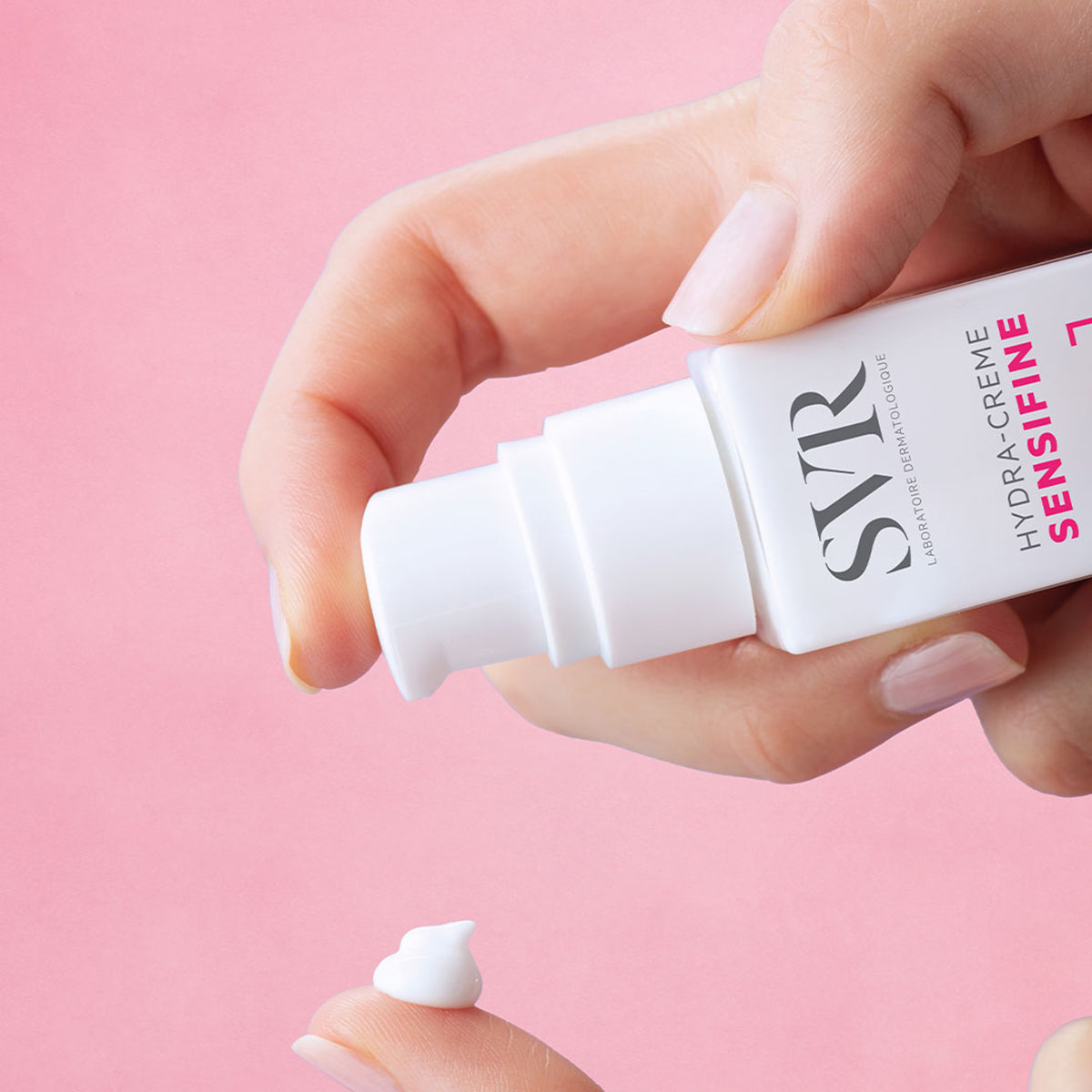 Mano aplicando crema hidratante blanca SVR Sensifine Hydra-Creme desde un envase dosificador blanco sobre fondo rosa.