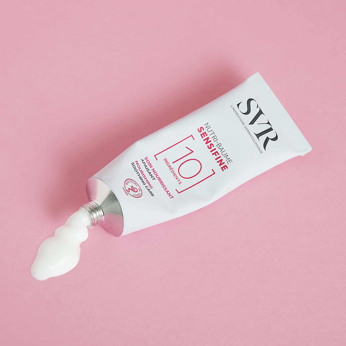 Tubo de aluminio SVR Sensifine Nutri-Baume con crema blanca saliendo sobre un fondo rosa suave.