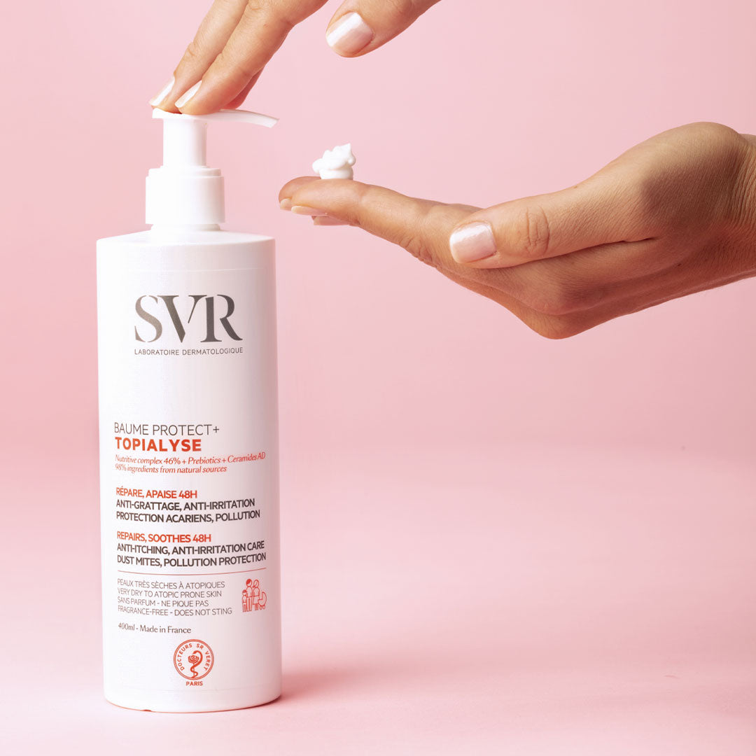 Bálsamo SVR TOPIALYSE Baume Protect+ en envase blanco con dosificador, aplicado sobre una mano contra fondo rosa.