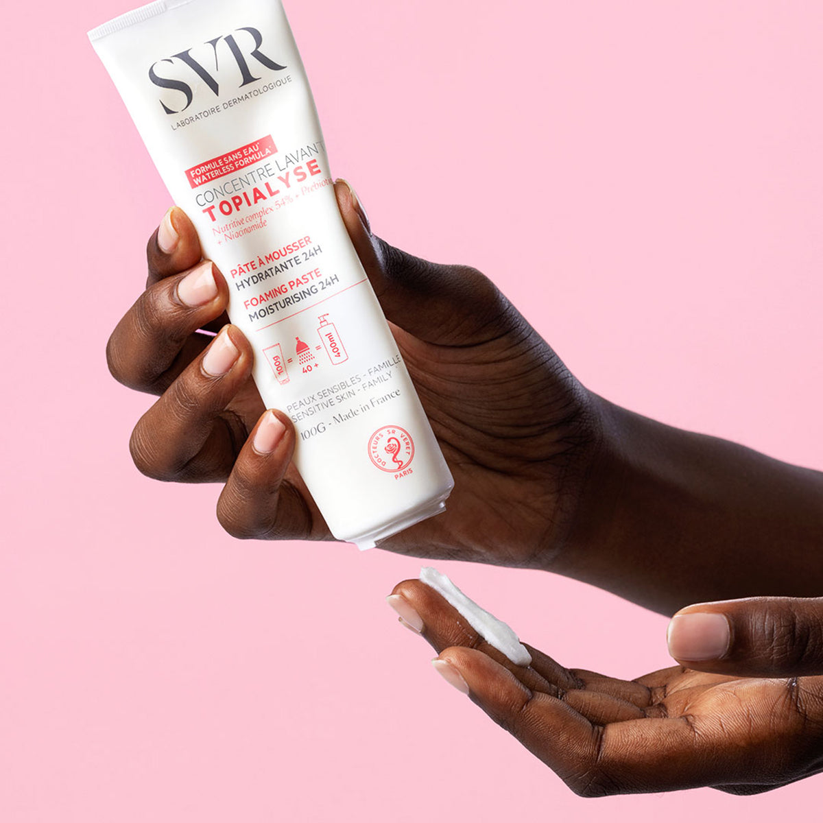 SVR Topialyse Concentré Lavant 100g tube being squeezed to dispense white foaming paste into a hand against a pink background.