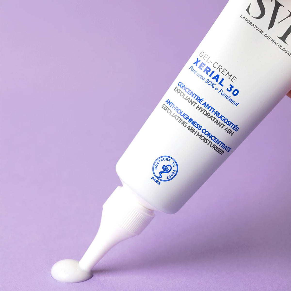 Tubo blanco de crema exfoliante SVR Xerial 30 dispensando una gota de producto sobre un fondo lila suave.