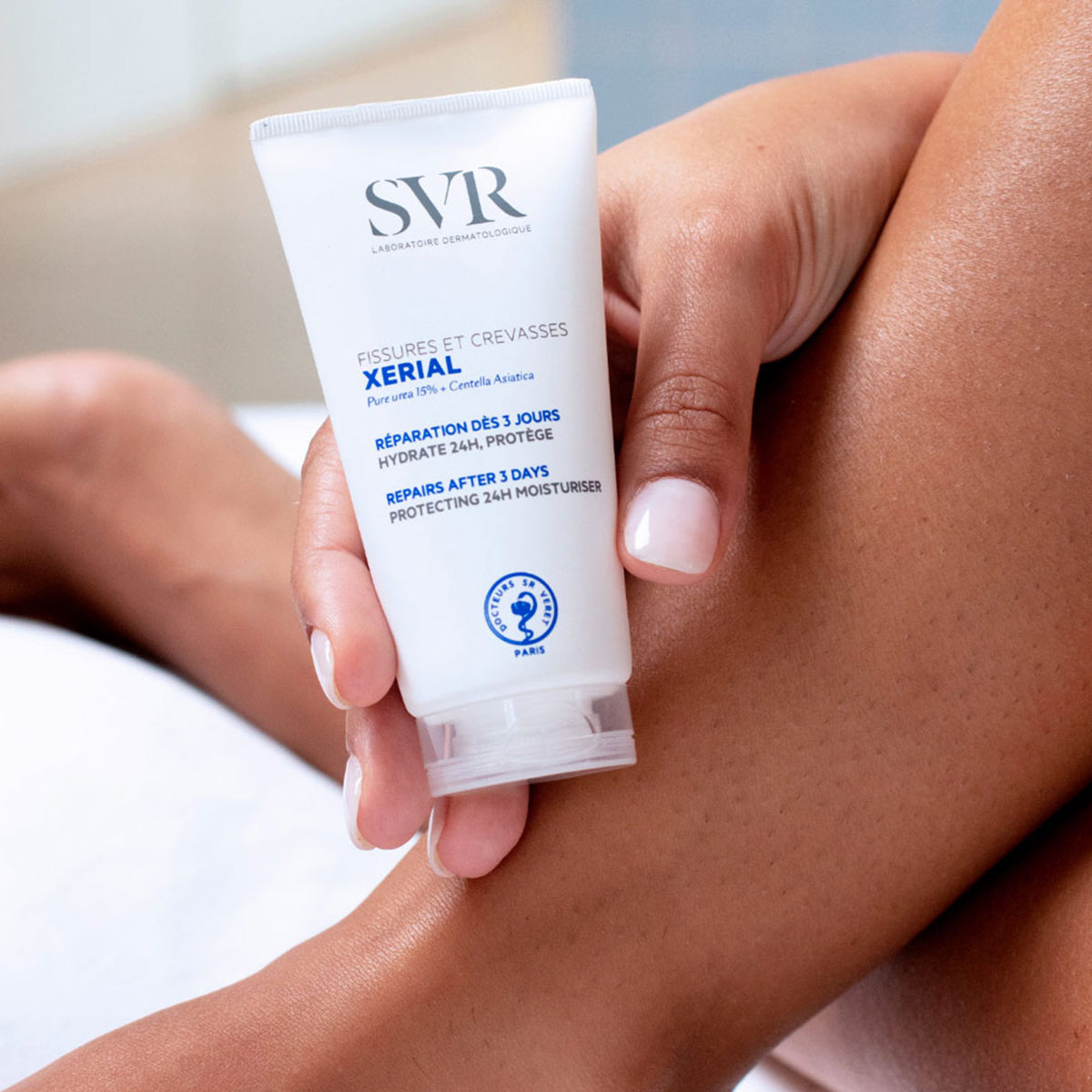 Tubo blanco de crema SVR Xerial Fisuras y Grietas sostenido en la mano sobre una pierna, diseño minimalista y profesional.