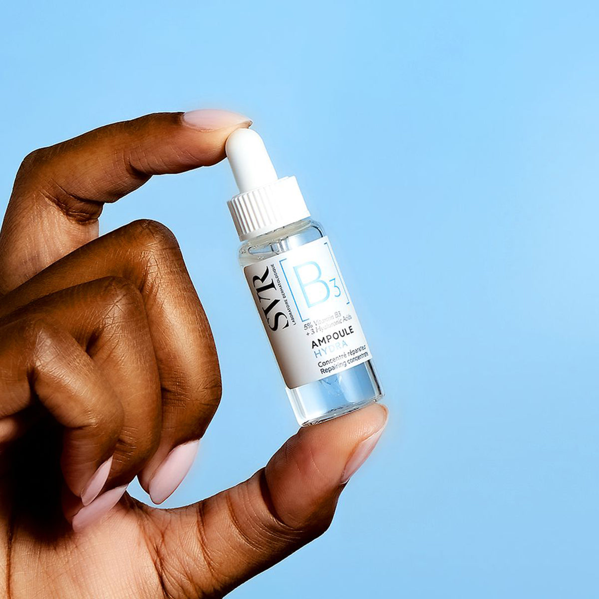 Mano sosteniendo un frasco de vidrio transparente SVR B3 Ampoule Hydra con gotero blanco sobre fondo azul claro.
