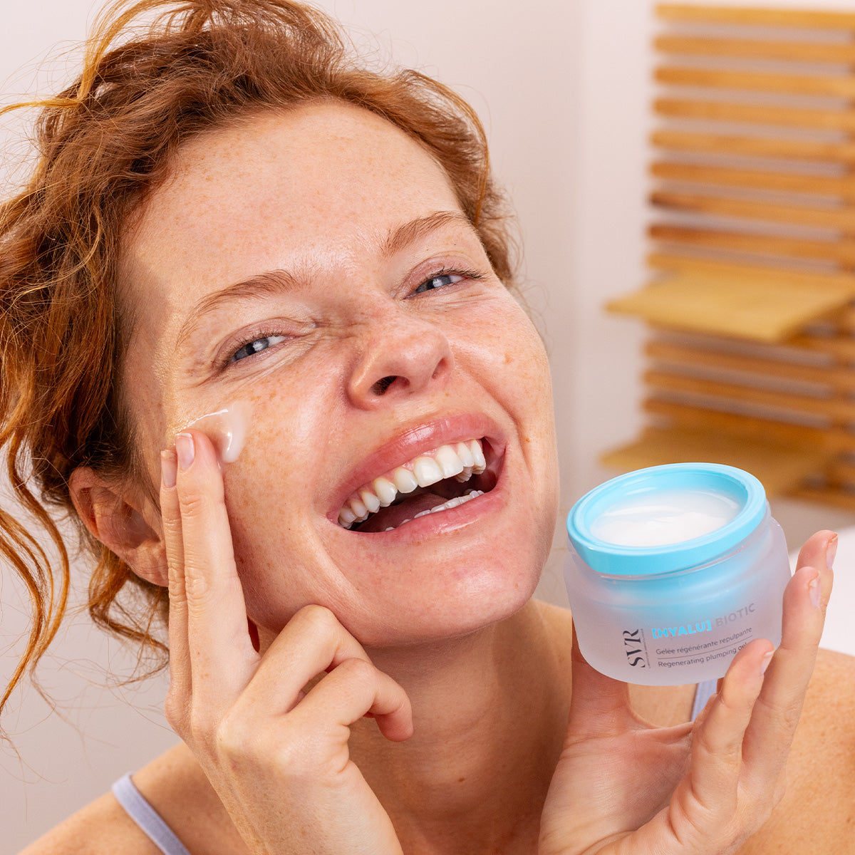 Mujer sonriente aplicando gel hidratante SVR Hyalu Biotic de tarro azul translúcido sobre su rostro.