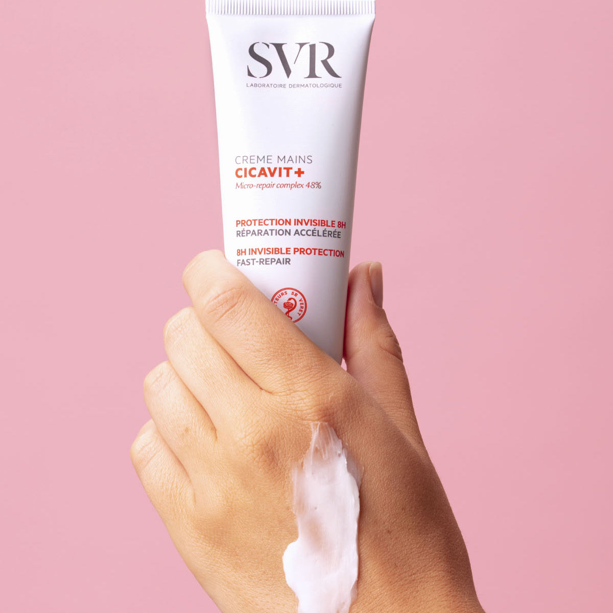 Tubo blanco de crema de manos SVR Cicavit+ sostenido en una mano con una muestra de textura cremosa sobre fondo rosa.