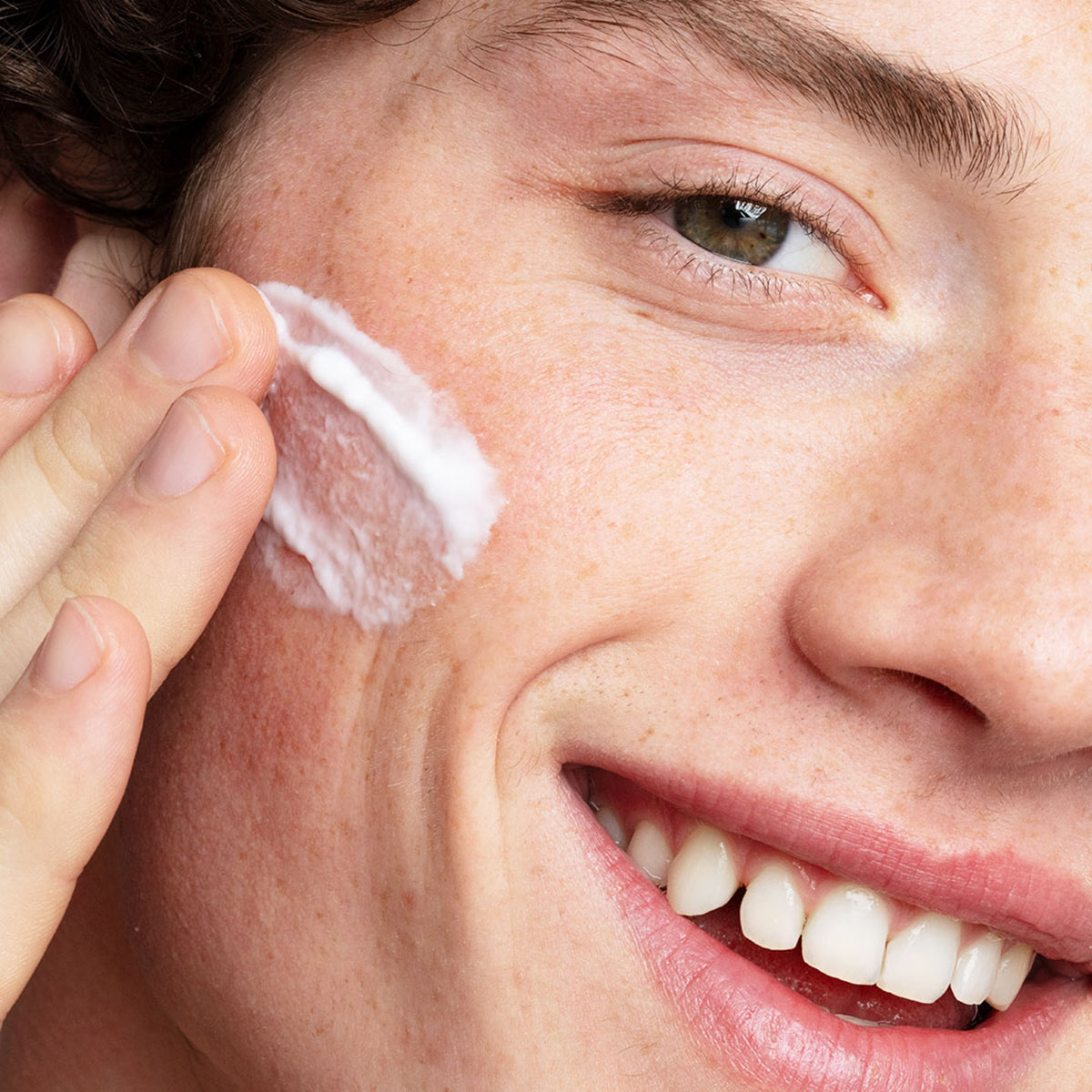 Persona sonriente aplicándose una crema facial blanca de textura ligera y fresca sobre la mejilla.