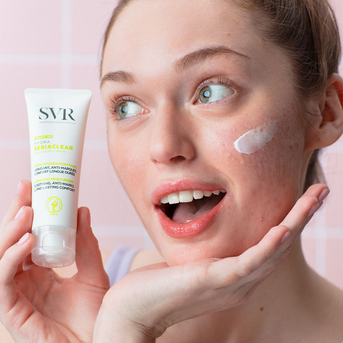 Mujer sosteniendo un tubo blanco de crema SVR Sebiaclear Hydra con una muestra de producto aplicada en su mejilla.
