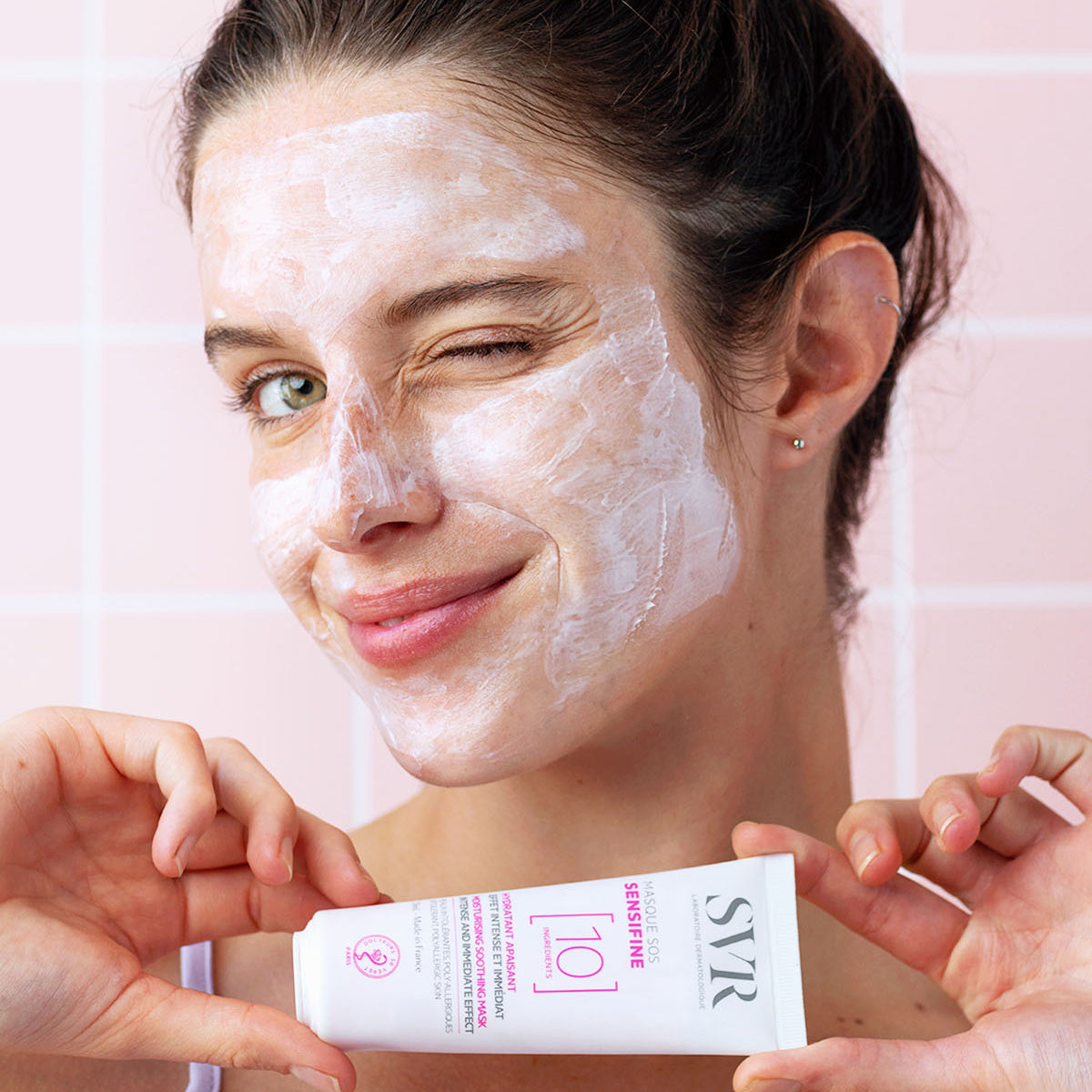 Woman winking with white SVR Sensifine SOS soothing face mask applied, holding the white product tube against a pink tile background.