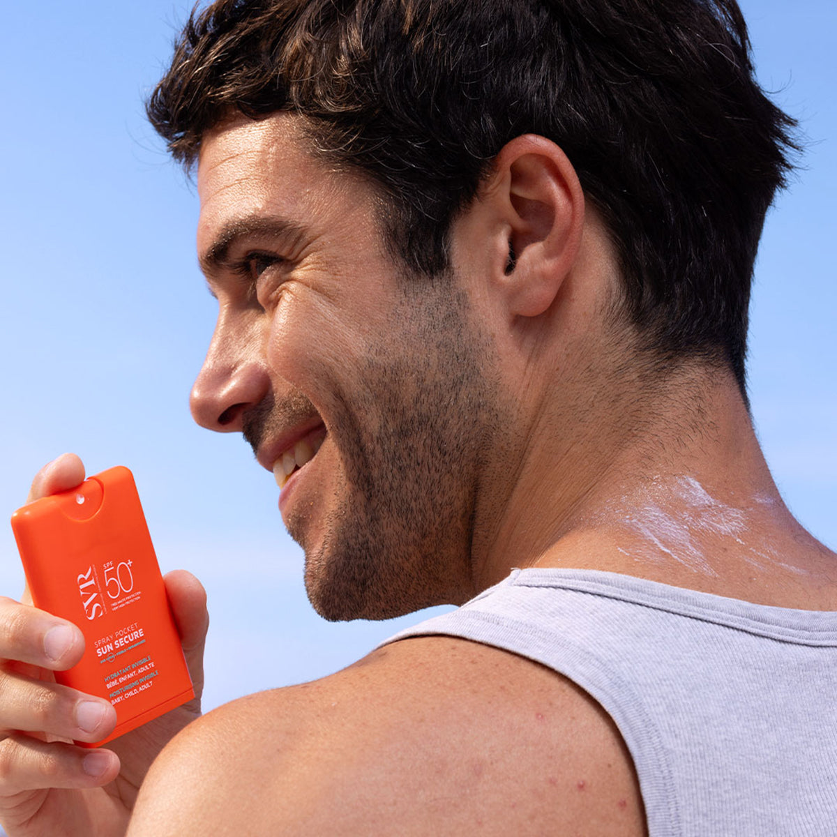Hombre sonriente aplicando protector solar SVR Sun Secure SPF50+ en formato spray naranja compacto sobre su cuello.