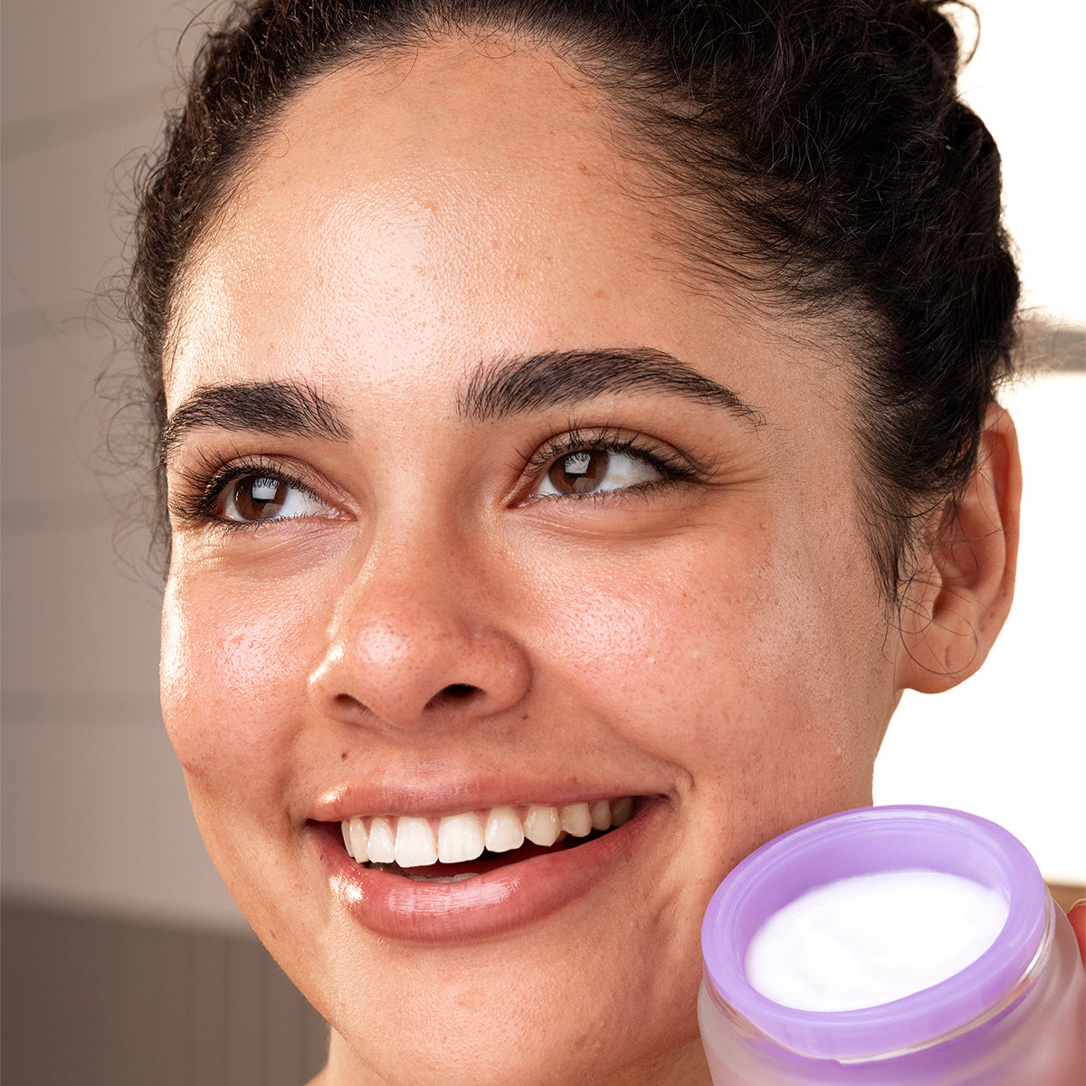 Mujer sonriente con piel radiante sosteniendo un frasco de crema facial PEPTI Biotic de tapa morada y textura cremosa.
