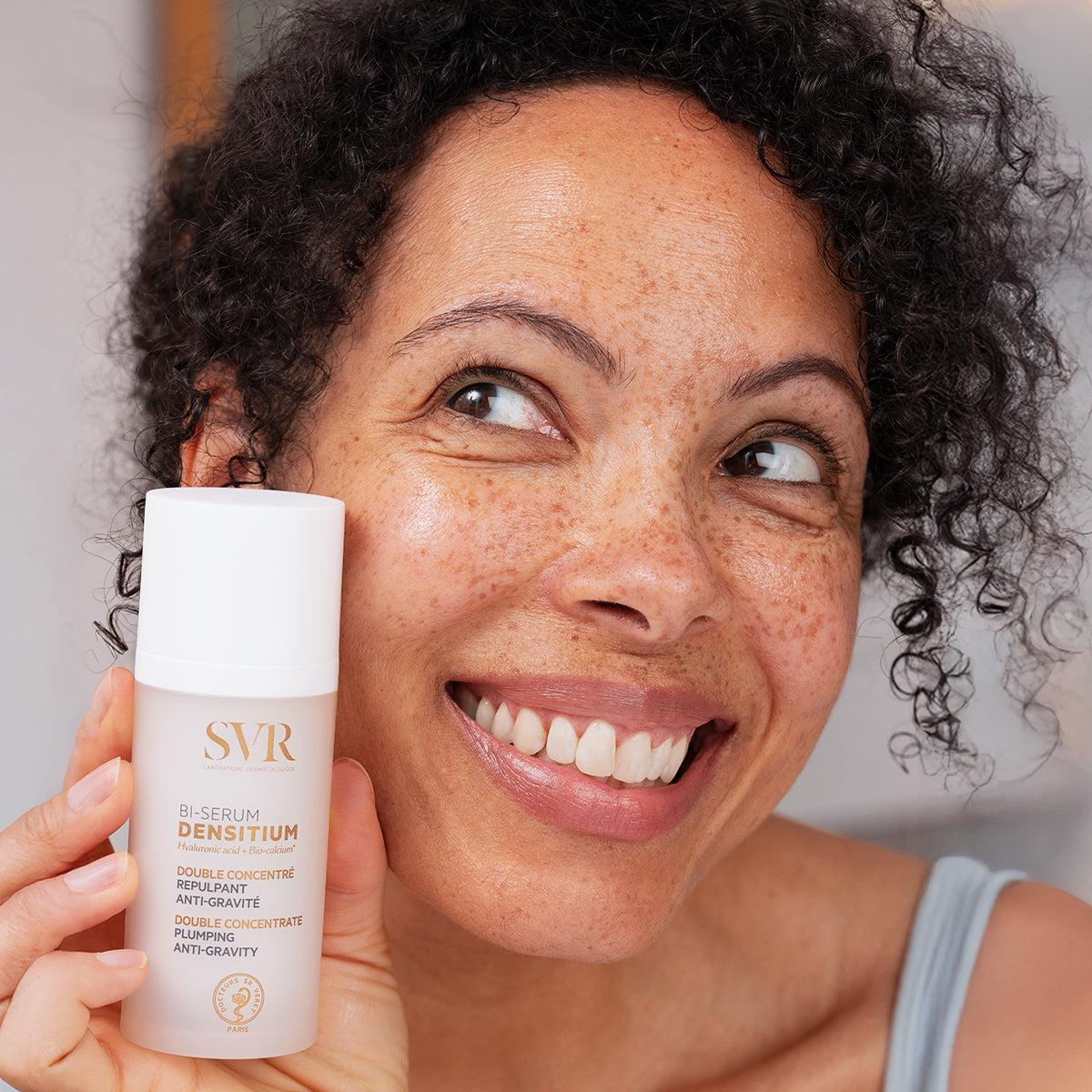 Mujer sonriente sosteniendo el frasco blanco y elegante de SVR Densitium Bi-Sérum antiedad para pieles maduras.