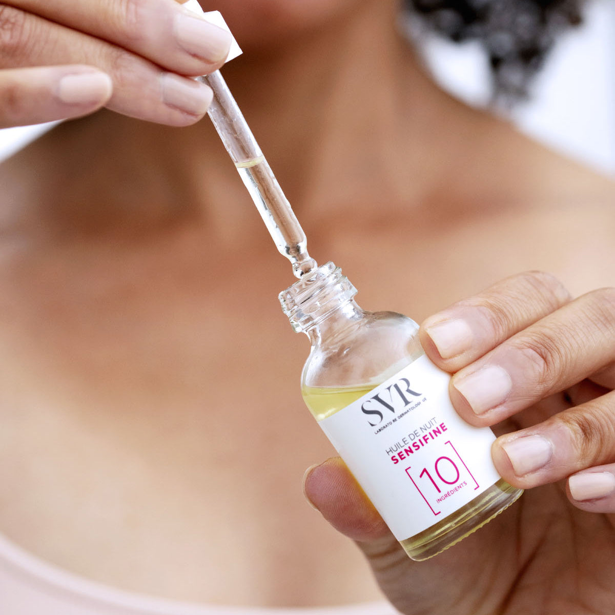Frasco de vidrio de aceite de noche SVR Sensifine con cuentagotas y etiqueta blanca minimalista en manos de una mujer.