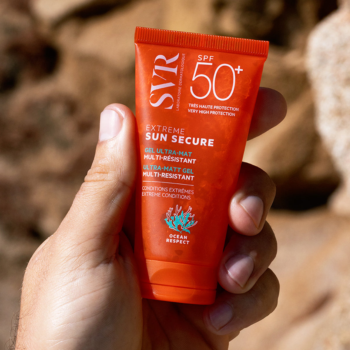 Tubo naranja de protector solar SVR Sun Secure Extreme SPF50+ en gel ultra mate sostenido en una mano sobre fondo rocoso.