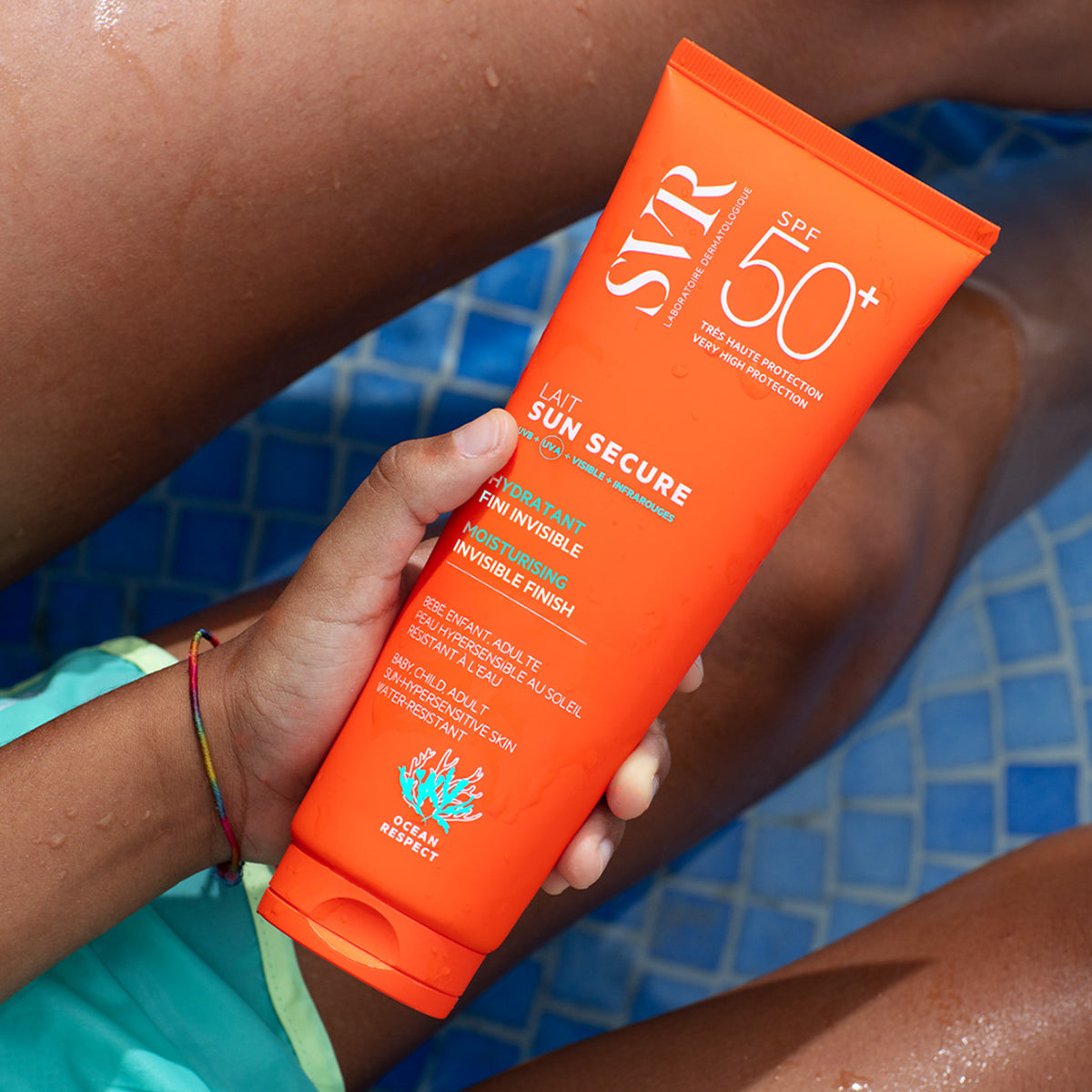 Tubo naranja de protector solar SVR Sun Secure SPF50+ sostenido por una mano sobre un fondo de piscina azul.