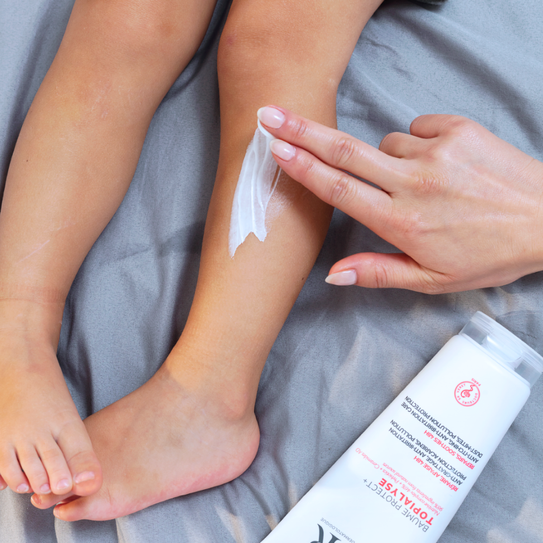 Mano aplicando bálsamo blanco TOPIALYSE Baume Protect+ sobre la pierna de un niño sobre una sábana gris.