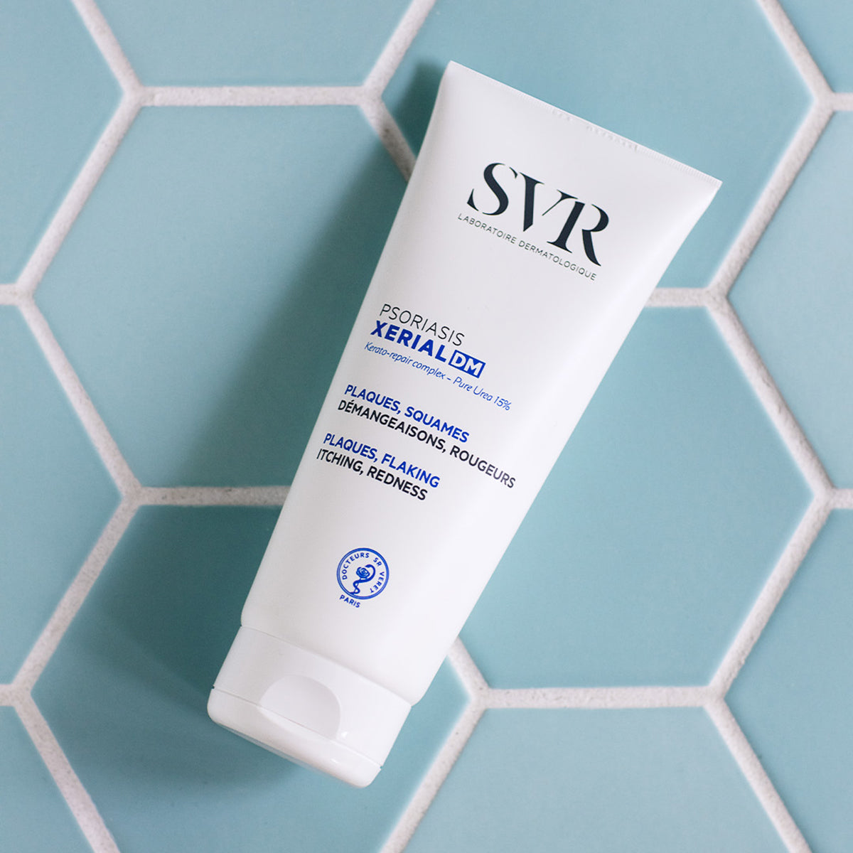 Tubo blanco de crema SVR XERIAL DM para psoriasis sobre un fondo de azulejos hexagonales de color azul claro.