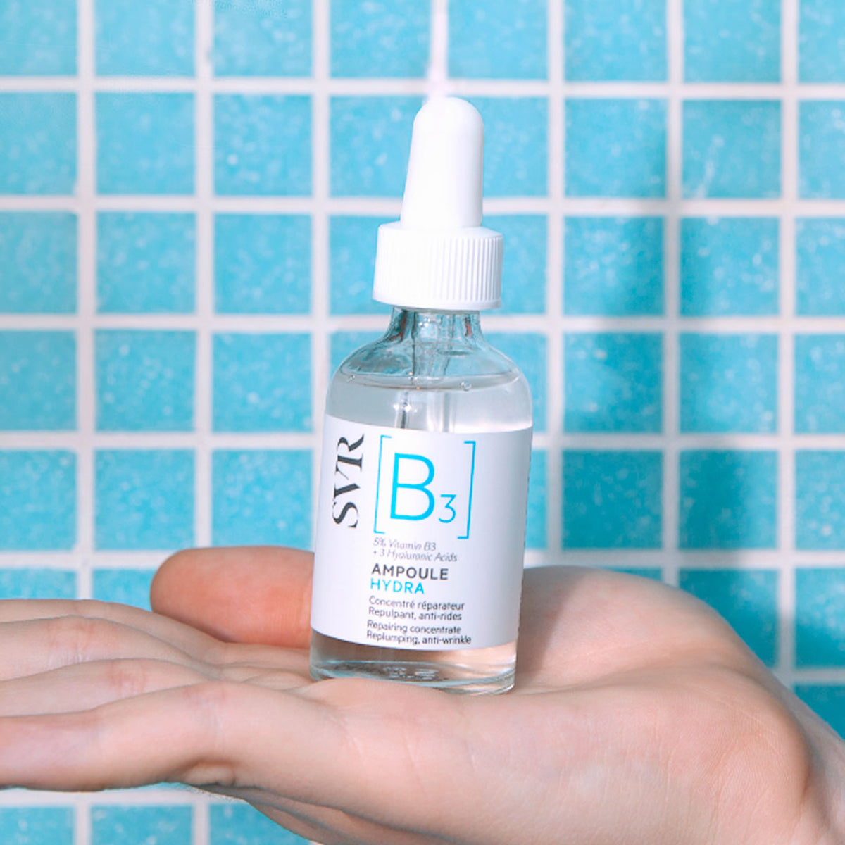 Frasco de cristal SVR Ampoule Hydra con gotero blanco sostenido en una mano frente a un fondo de azulejos azules.