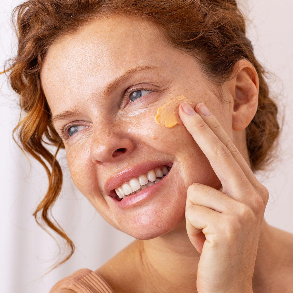 Mujer pelirroja aplicándose crema solar SUN SECURE Blur SPF50+ de textura mousse color albaricoque en la mejilla.