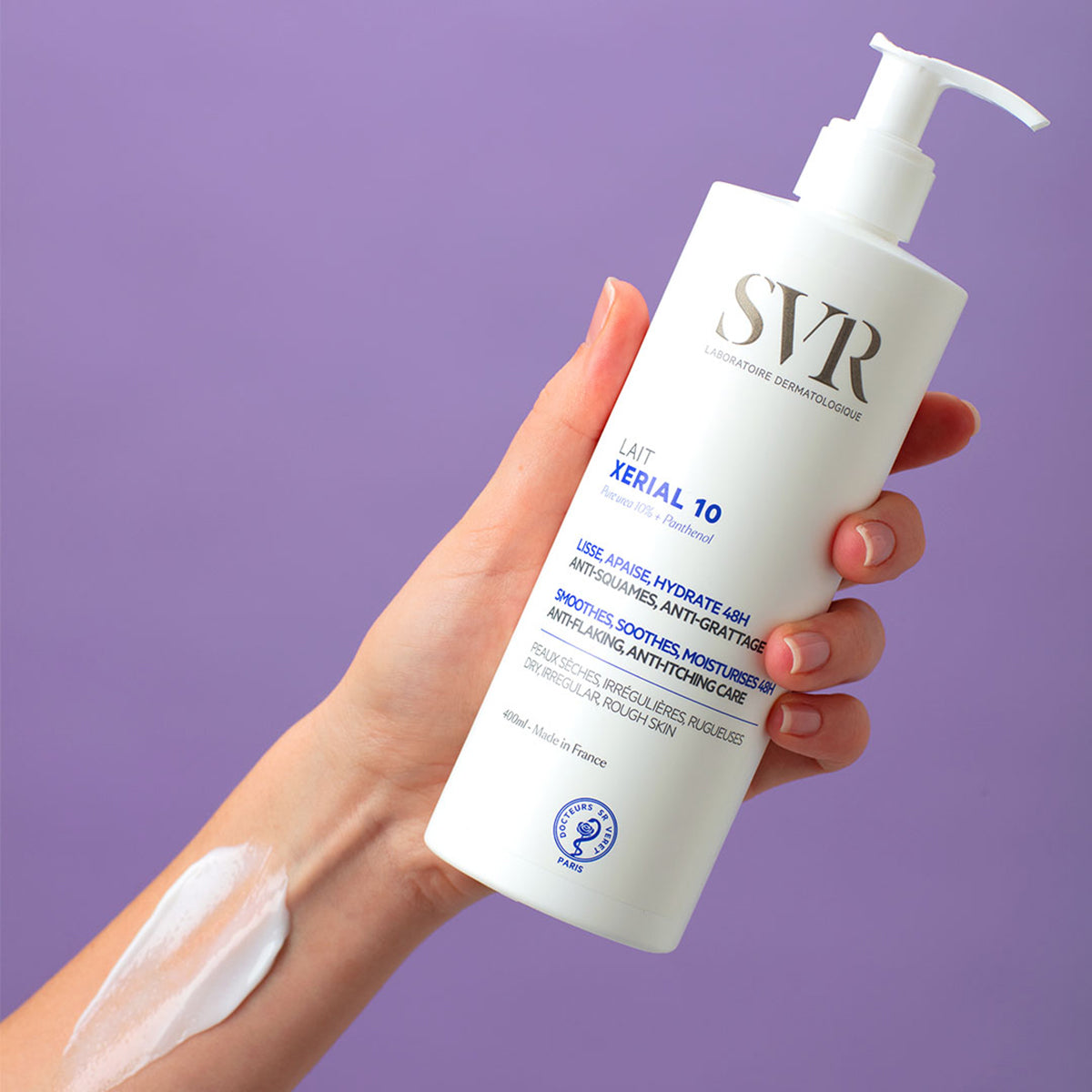 Mano sosteniendo un frasco blanco de loción corporal SVR Xerial 10 de 400ml con textura cremosa sobre fondo morado.