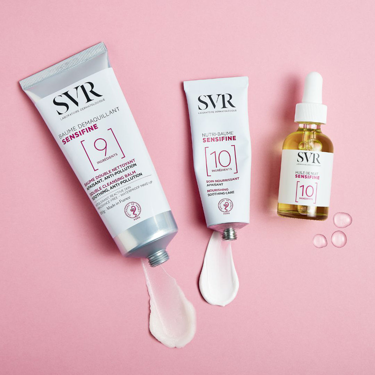 Dos tubos de aluminio SVR Sensifine y un frasco de aceite sobre fondo rosa con muestras de crema blanca.