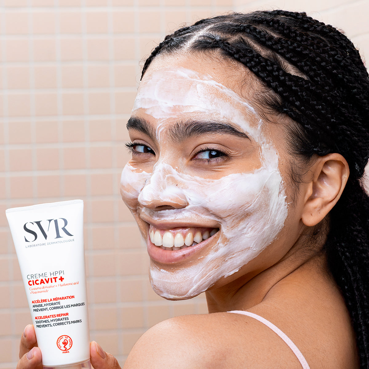 Mujer sonriente con crema reparadora blanca SVR Cicavit+ HPPI aplicada en el rostro sobre un fondo de azulejos beige.