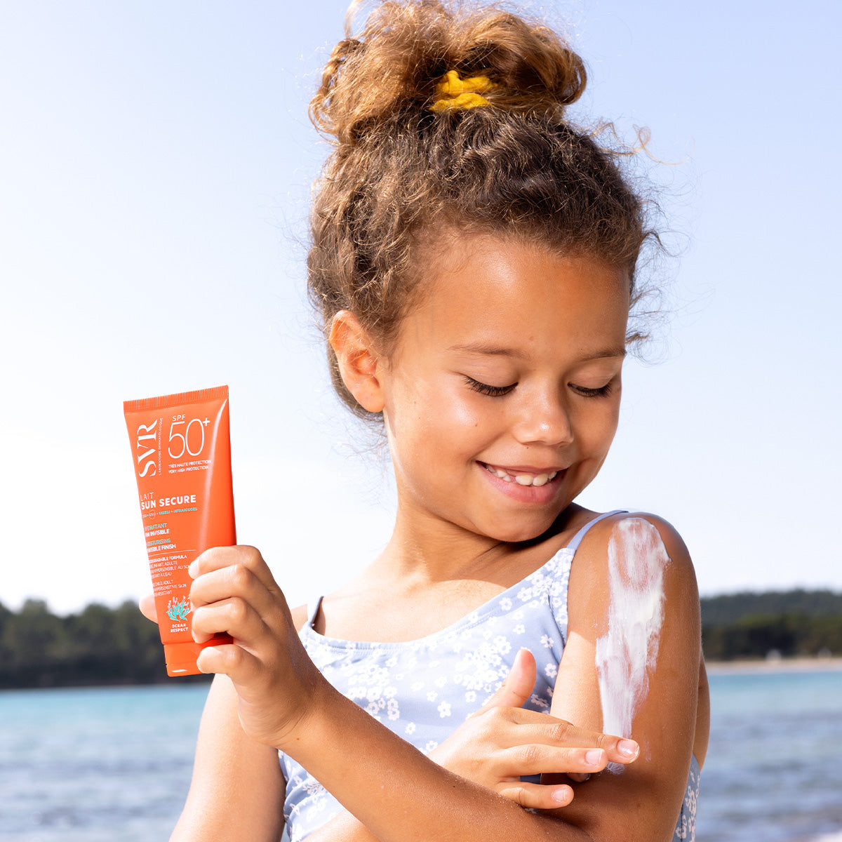 A young girl applying SVR SUN SECURE SPF50+ sunscreen lotion to her shoulder on a sunny beach.