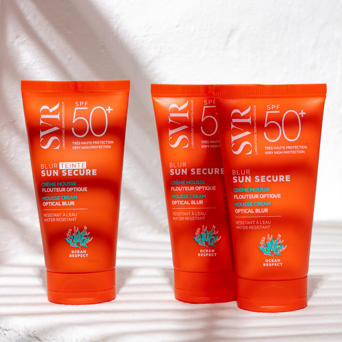 Tres tubos naranjas de protector solar SVR Sun Secure Blur Teinte SPF50+ con textura mousse sobre fondo blanco.