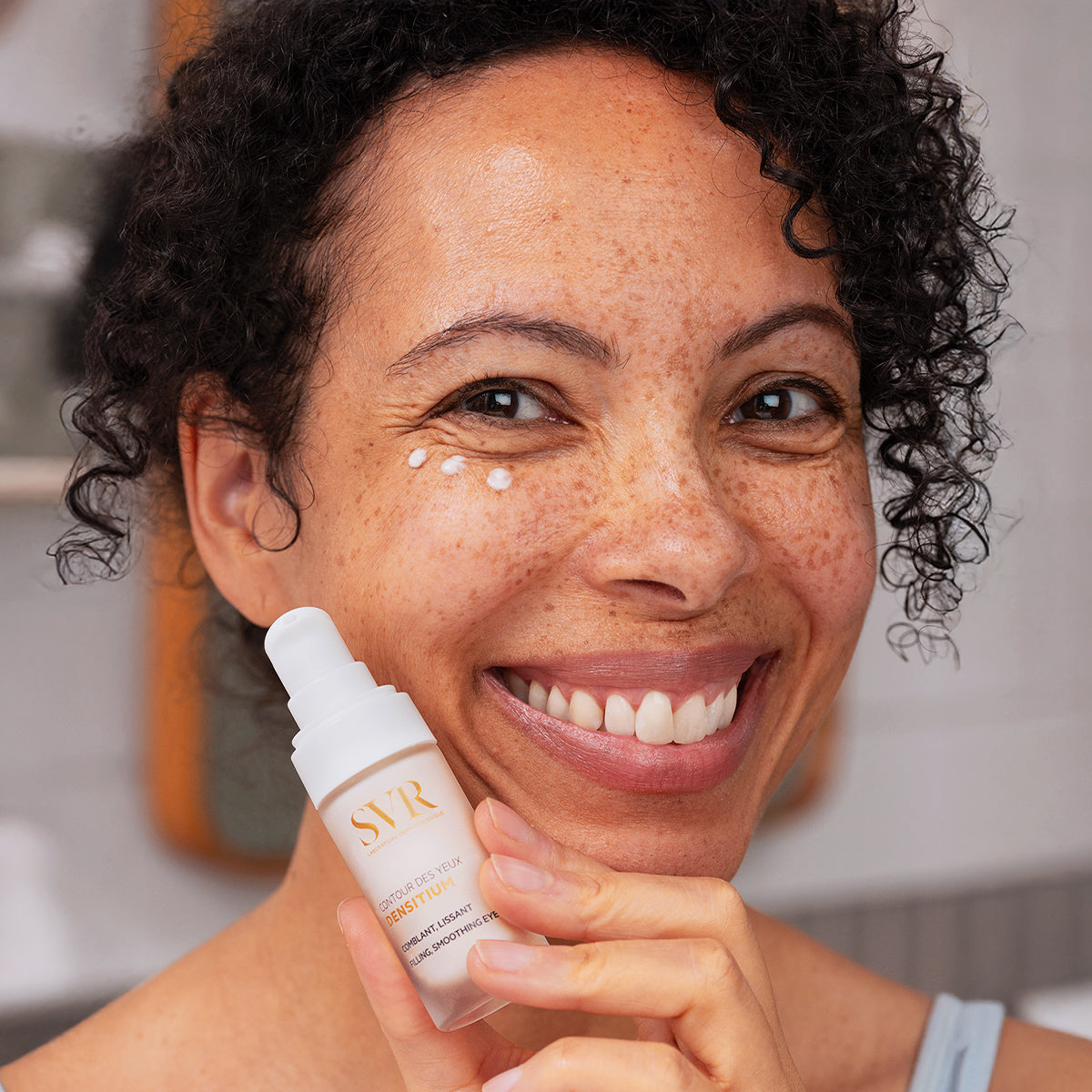 Mujer sonriente aplicando crema SVR Densitium Contour des Yeux en el contorno de ojos con puntos de producto blanco.