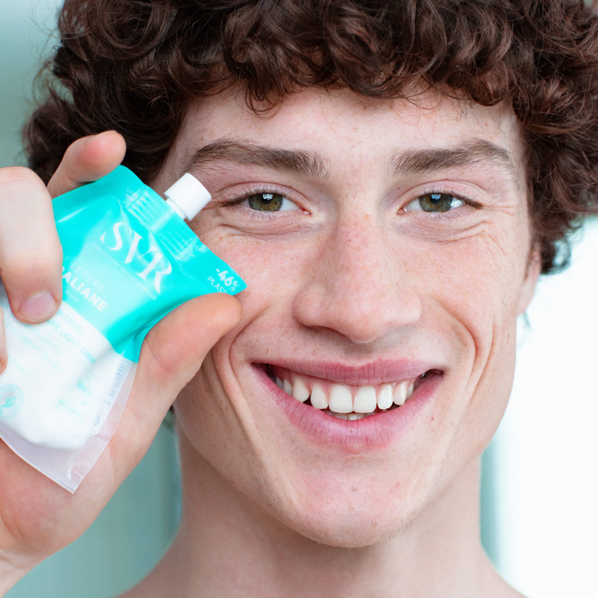 Joven sonriente sosteniendo un envase flexible de crema hidratante SVR Hydraliane color turquesa cerca de su rostro.