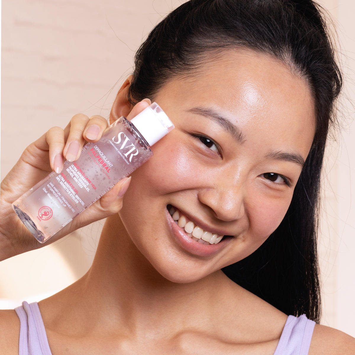 Mujer sonriente sosteniendo un frasco transparente de gel desmaquillante SVR Palpebral para ojos sensibles.