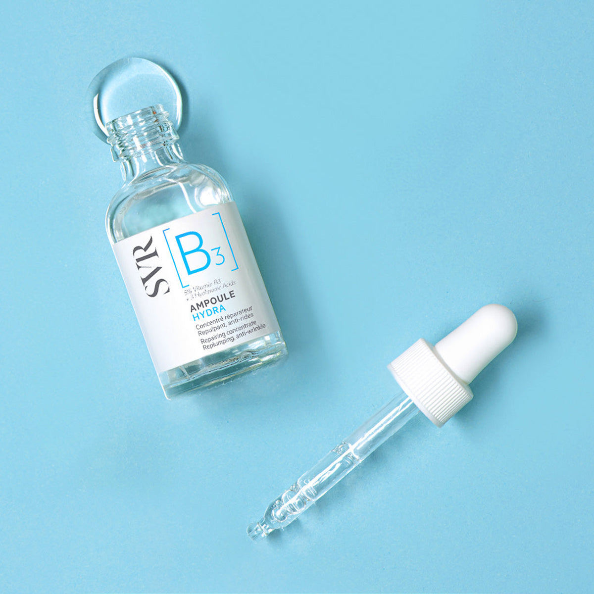 Frasco de vidrio transparente SVR Ampoule Hydra con vitamina B3 y gotero blanco sobre fondo azul claro.