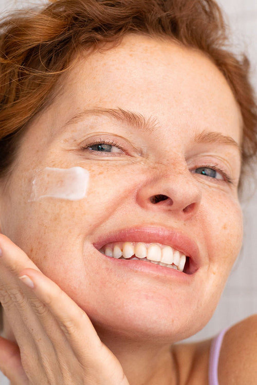 Mujer pelirroja con pecas aplicando crema facial blanca en su mejilla con una sonrisa radiante y natural.