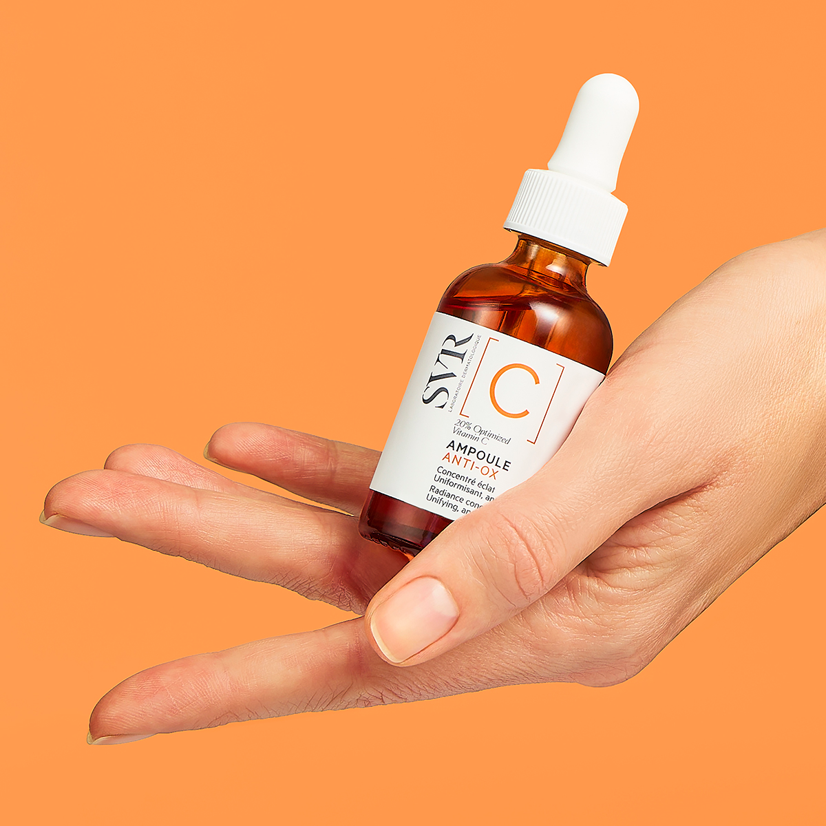Frasco de vidrio ámbar con gotero de SVR Ampoule Anti-Ox sostenido en una mano sobre fondo naranja vibrante.