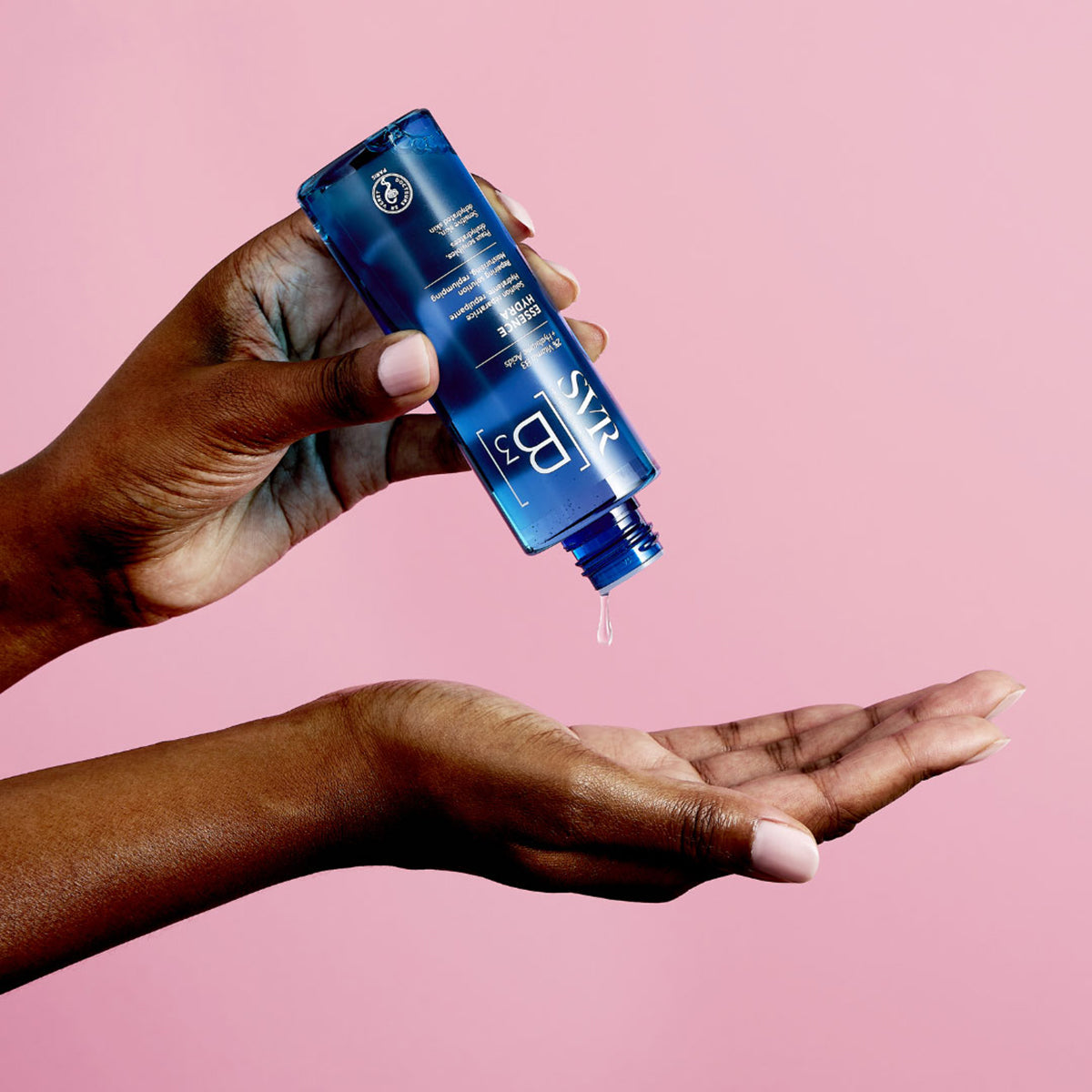 Frasco azul de SVR B3 Essence Hydra vertiendo una gota de esencia hidratante sobre una mano contra fondo rosa.