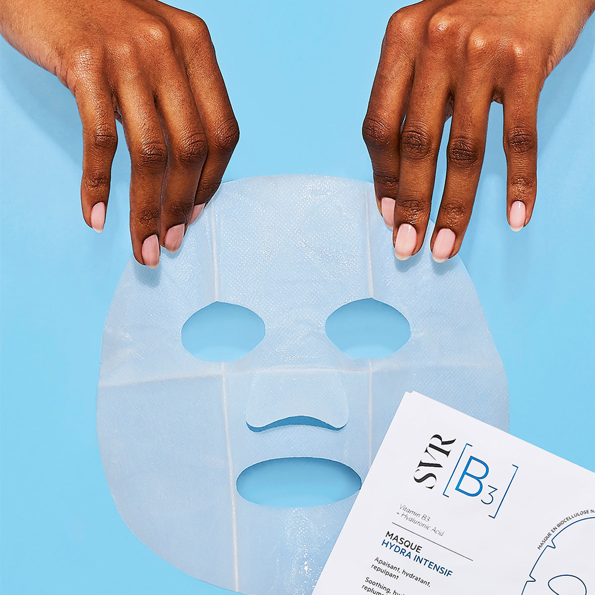 Manos sosteniendo una mascarilla facial de biocelulosa translúcida sobre un fondo azul junto al empaque SVR B3 Hydra Intensif.
