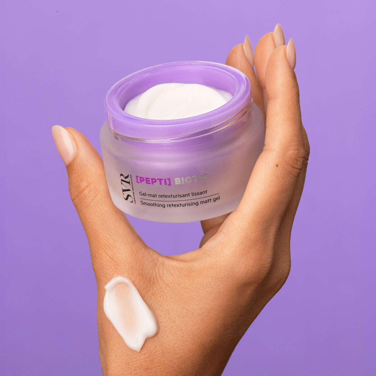 Tarro de gel regenerador SVR [PEPTI] Biotic con tapa lila y textura cremosa sobre una mano, fondo morado.