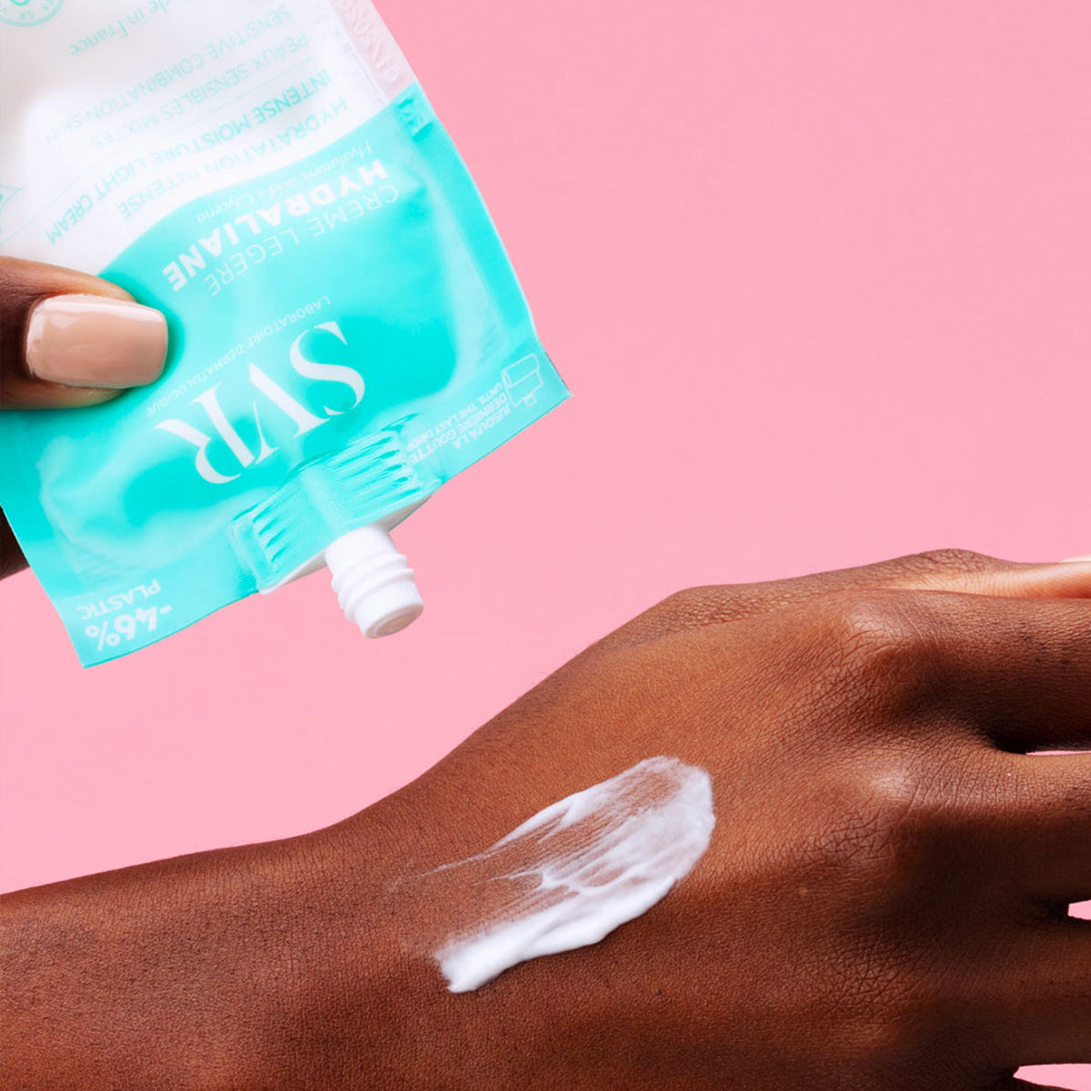 Envase flexible color turquesa de Hydraliane Légère aplicando crema hidratante blanca sobre una mano contra fondo rosa.