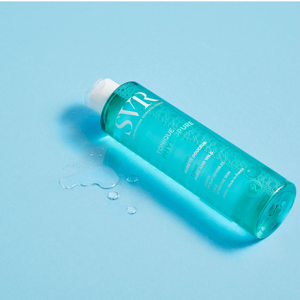 Frasco de 200ml de tónico facial SVR PHYSIOPURE transparente y azul sobre fondo celeste con gotas de producto.