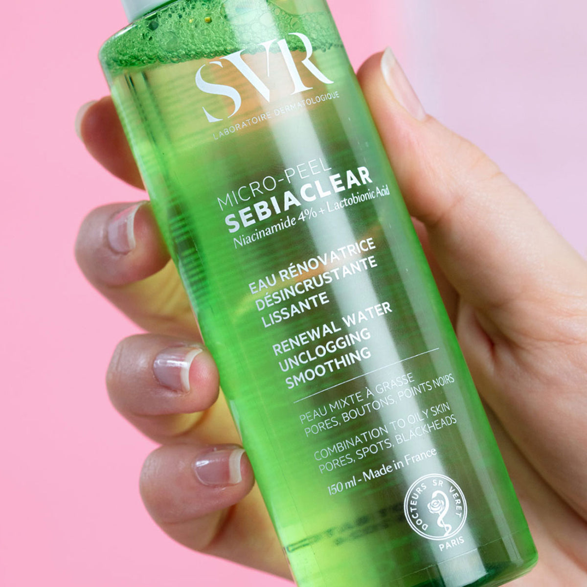 Frasco transparente de 150 ml con líquido verde de SVR Sebiaclear Micro-peel sostenido en una mano sobre fondo rosa.