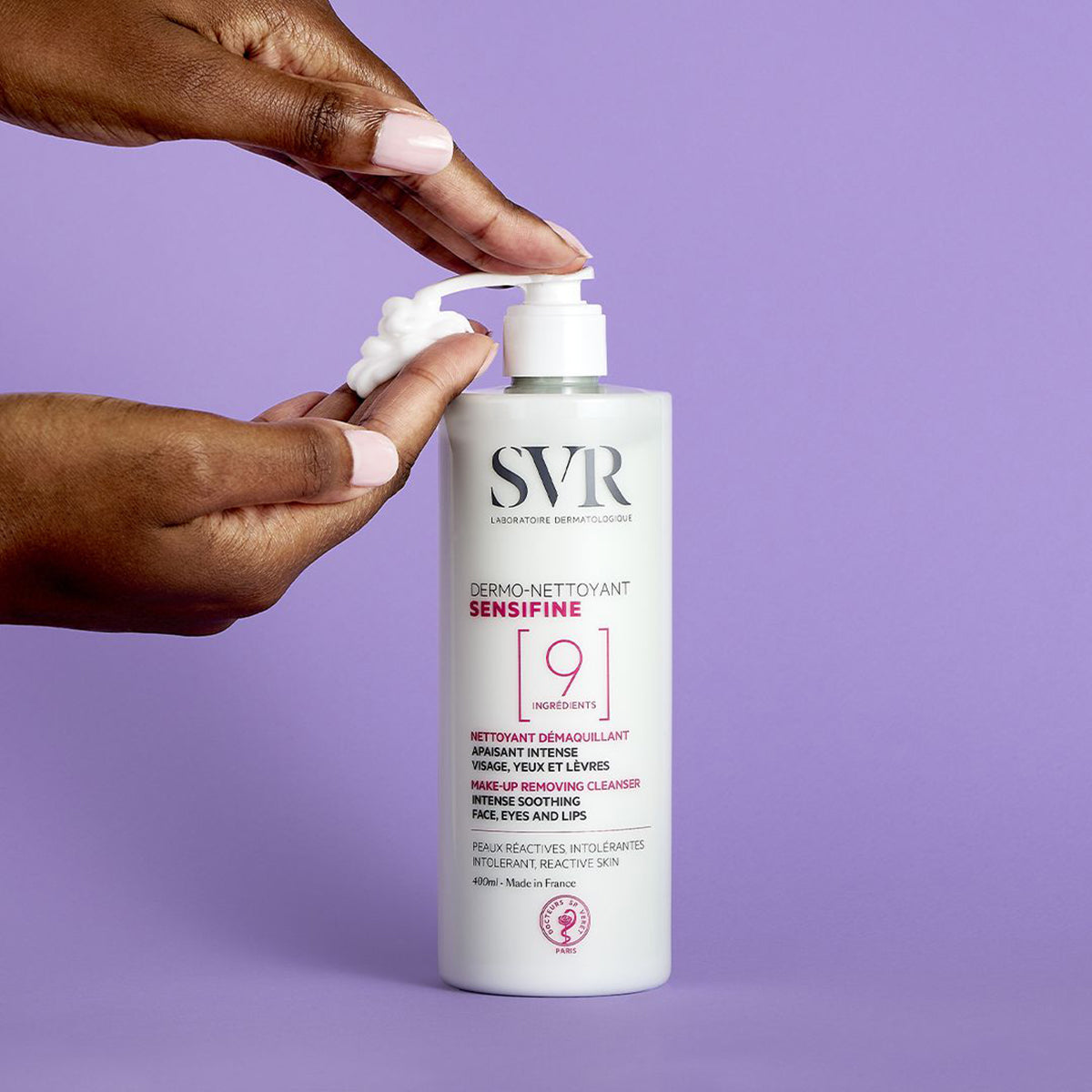 Botella blanca de SVR Sensifine Dermo-Nettoyant de 400ml con dosificador, siendo aplicada sobre una mano contra fondo morado.
