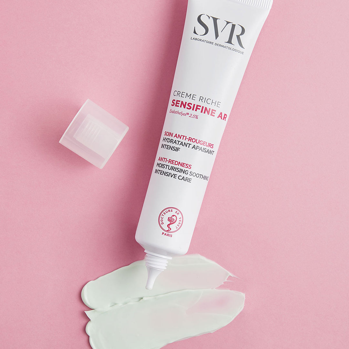 Tubo blanco de crema SVR Sensifine AR con tapa abierta y muestra de textura verde sobre fondo rosa.