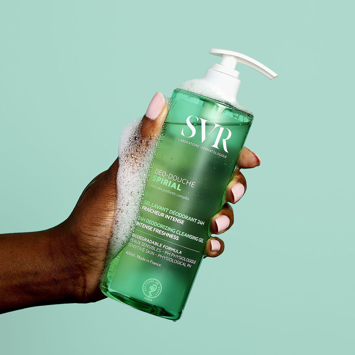 Mano sosteniendo un envase de 400ml de gel de ducha SVR Spirial verde con dosificador y espuma sobre fondo verde claro.