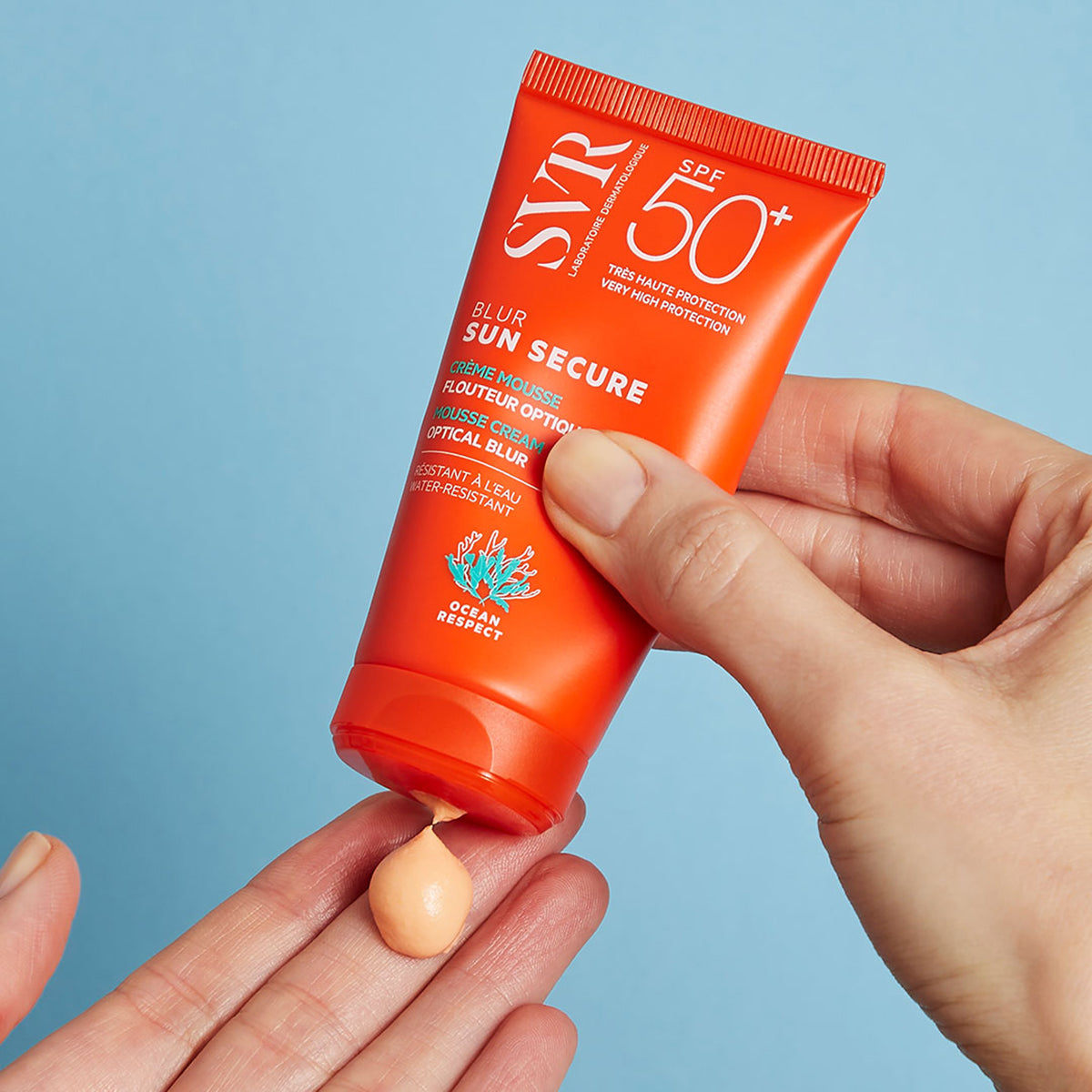 Tubo naranja de protector solar SVR SUN SECURE Blur SPF50+ dispensando una crema mousse color albaricoque sobre una mano.