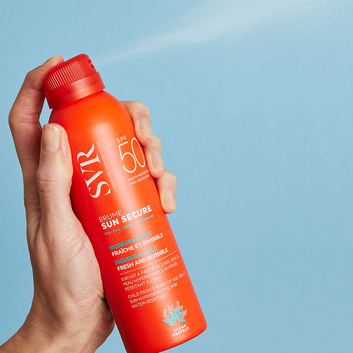 Mano sosteniendo un envase naranja de protector solar SVR Sun Secure Brume SPF50 con pulverizador sobre fondo azul.