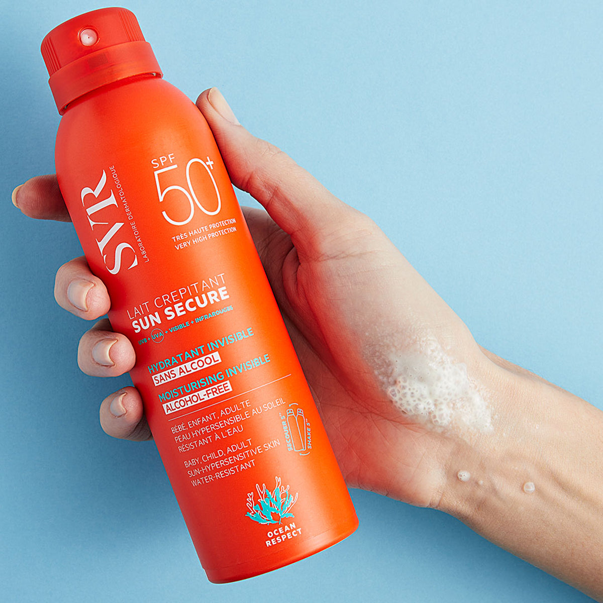 Mano sosteniendo un spray solar naranja SVR Sun Secure SPF50+ con textura espumosa sobre la piel contra un fondo azul.
