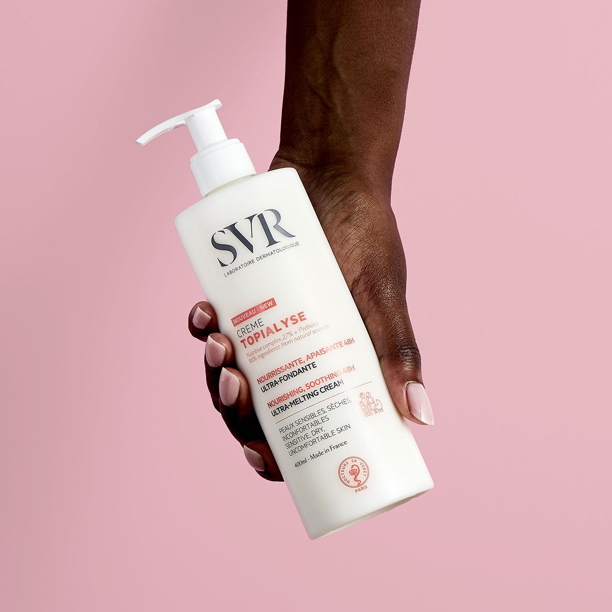 Mano sosteniendo un frasco blanco de 400ml de crema hidratante SVR Topialyse con dosificador sobre fondo rosa.